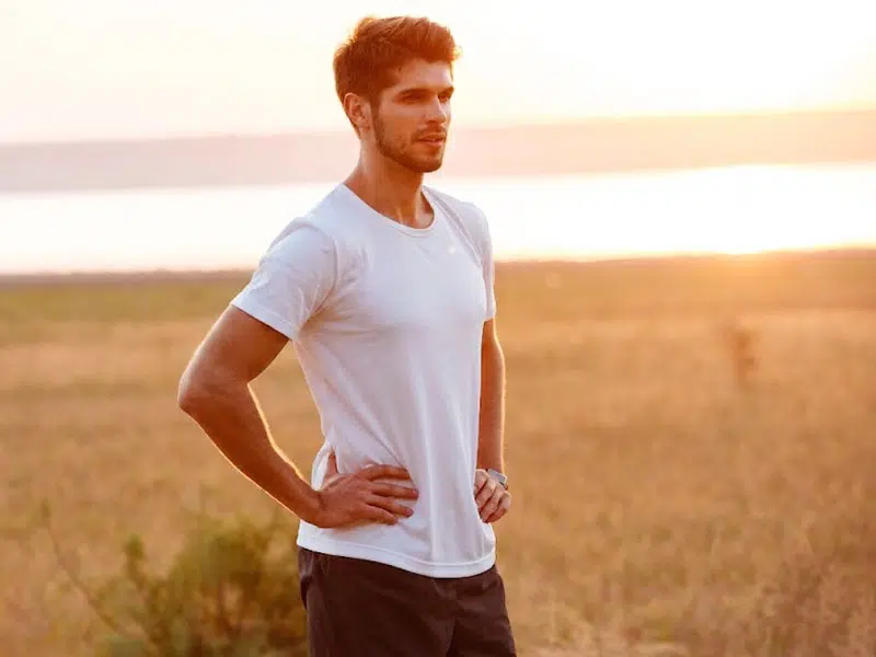 Sportlicher Mann im weißen T-Shirt steht bei Sonnenaufgang in einer Feldlandschaft am Wasser. © vadymvdrobot, 123rf.com.