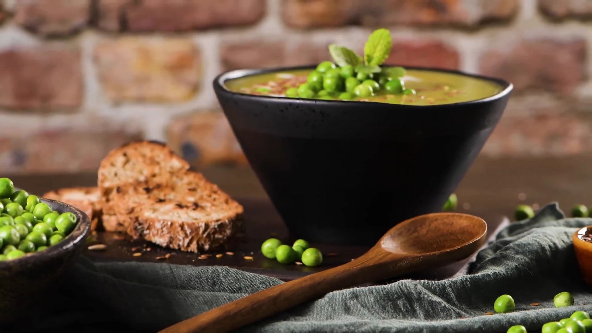 Erbsensuppe in schwarzer Schale, daneben Brot und frische Erbsen